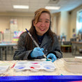 Load image into Gallery viewer, Artist Andrea S. smiling as she dips her paintbrush into green paint